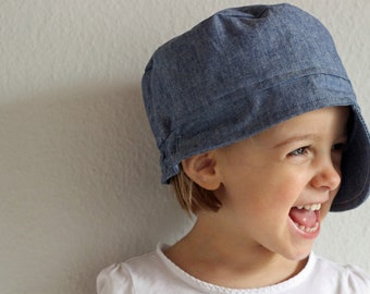 Op de afbeelding: Een jong kind draagt een blauwe denim newsboy-pet met de rand omhoog gevouwen aan één kant. Het kind glimlacht en kijkt naar rechts.