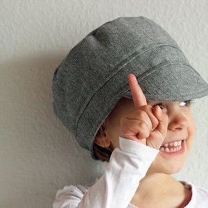 Puede incluir: Un niño pequeño con una gorra de newsboy gris sonríe y señala con el dedo índice. El niño lleva una camisa blanca de manga larga.