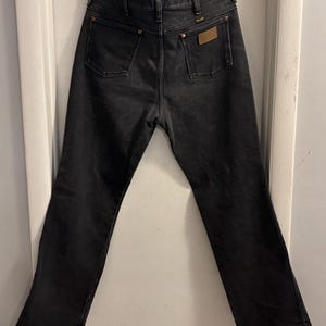 May include: Black denim jeans with two back pockets and a small brown patch. The jeans have a classic five-pocket design and a straight leg fit. The jeans are hanging on a white door.