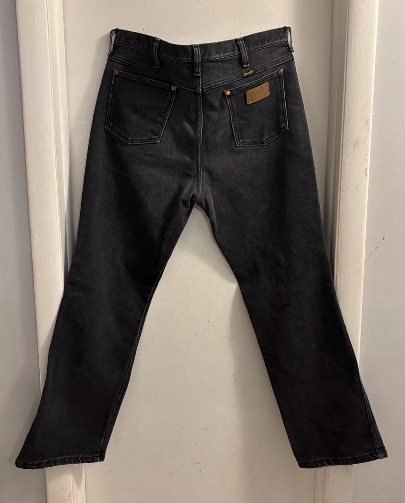May include: Black denim jeans with two back pockets and a small brown patch. The jeans have a classic five-pocket design and a straight leg fit. The jeans are hanging on a white door.