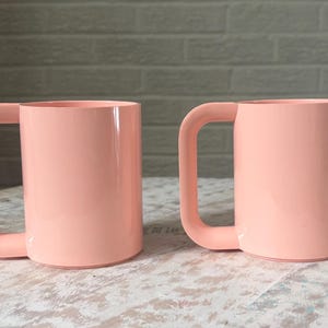 May include: Two peach-colored mugs with large, square handles sit side-by-side on a distressed white table. The mugs have a smooth, glossy finish and a cylindrical shape. The background features a light gray brick wall.