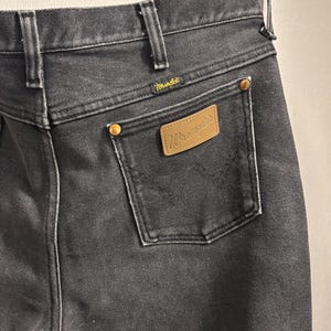 May include: Close-up of black denim jeans with a Wrangler label on the back pocket. The jeans have a classic five-pocket design, belt loops, and brass-coloured rivets. The Wrangler logo is visible on the label and above the back pocket.