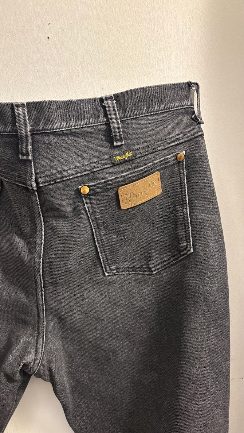May include: Close-up of black denim jeans with a Wrangler label on the back pocket. The jeans have a classic five-pocket design, belt loops, and brass-coloured rivets. The Wrangler logo is visible on the label and above the back pocket.