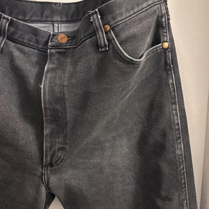 May include: Close-up of black denim jeans. The jeans have a classic five-pocket design with a button and zip closure. The jeans are made of a durable denim fabric and feature copper-coloured rivets.