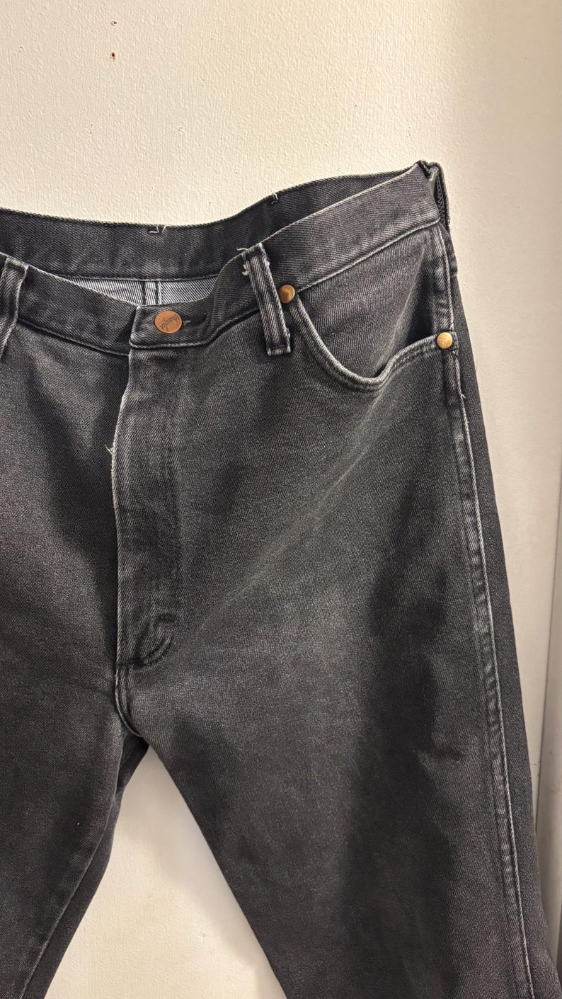 May include: Close-up of black denim jeans. The jeans have a classic five-pocket design with a button and zip closure. The jeans are made of a durable denim fabric and feature copper-coloured rivets.