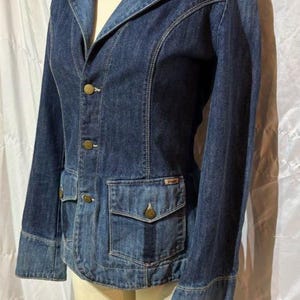 May include: A dark blue denim jacket featuring a notched lapel, long sleeves, and a button-front closure. The jacket includes two front patch pockets with buttoned flaps and visible topstitching details throughout the garment structure.