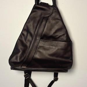 May include: A dark brown leather backpack with a triangular shape. It has a top handle, a front pocket, and adjustable shoulder straps with metal buckles. The bag is hanging against a white wall.