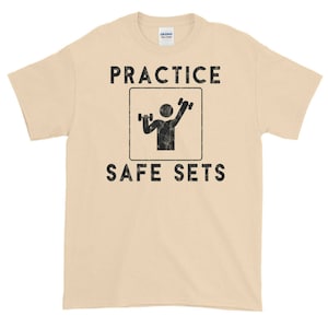 Könnte beinhalten: Ein beiges T-Shirt mit einer schwarzen Grafik einer Person, die Gewichte hebt, in einem Schild mit der Aufschrift "Practice Safe Sets".