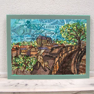Può includere: Un'opera d'arte in collage incorniciata che presenta una città con edifici, un ponte e un albero. L'opera è realizzata con ritagli di carta e presenta una combinazione di colori blu, verde e marrone. La parola "LEIDEN" è visibile nell'opera.