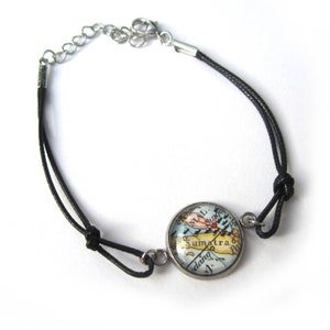 Op de afbeelding: Een armband van zwart koord met een zilveren sluiting en een ronde glazen bedel met een vintage kaart van Sumatra. De kaart is in tinten blauw, groen en bruin met rode markeringen.