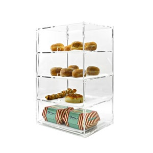 May include: A clear acrylic display case with four shelves, filled with pastries and snacks. The case has a white background and is on a white surface. The pastries include doughnuts, bread rolls, and a pastry with a swirl design. The case also contains two boxes with the word "Yumm!" printed on them.