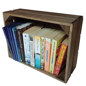May include: A wooden crate bookshelf, filled with a selection of books. The books have different colored spines and titles such as "The Last Fighting Tommy" and "Island of Secrets". The crate is made of aged, brown wood.
