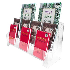 May include: A clear acrylic display stand with multiple shelves holding various 2021 diaries. The diaries have different covers, including a green and gold floral design, a red cover with the text "Progress not perfection", and a red cover with the text "2021 Diary".