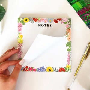 May include: A notepad with a floral border and the word "NOTES" printed in black. The notepad is white with a floral border in shades of pink, red, yellow, blue, and green. The notepad is partially open, revealing a blank white page.