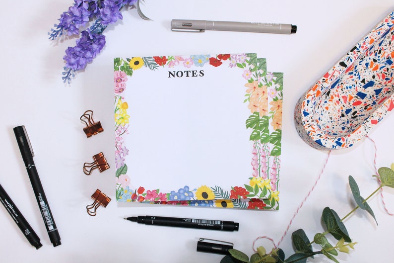 May include: A floral notepad with a white background and a black border. The notepad has the word "NOTES" printed in black at the top. The notepad is surrounded by a variety of colorful flowers, including pink, yellow, blue, and red.