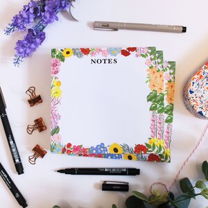May include: A white notepad with a floral border and the word "NOTES" printed in black. The notepad is surrounded by pens, clips, and other office supplies.