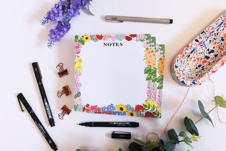 May include: A white notepad with a floral border and the word "NOTES" printed in black. The notepad is surrounded by pens, clips, and other office supplies.