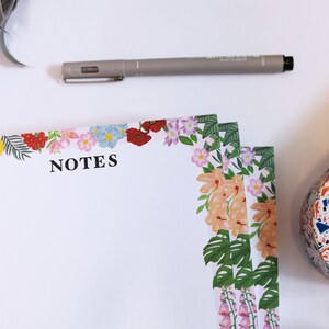 May include: A stack of floral notepad paper with the word "NOTES" printed on the top. The paper has a colorful floral border with pink, yellow, red, and green flowers.