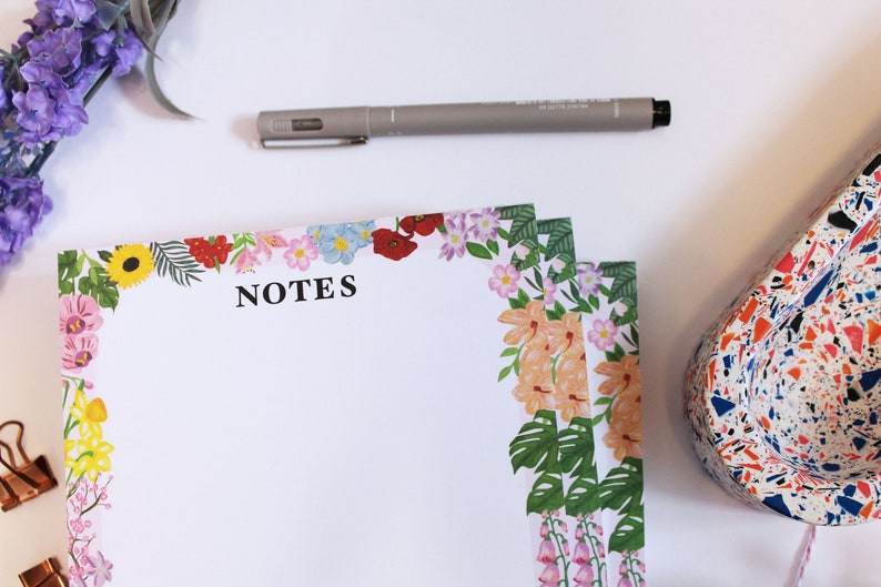 May include: A stack of floral notepad paper with the word "NOTES" printed on the top. The paper has a colorful floral border with pink, yellow, red, and green flowers.