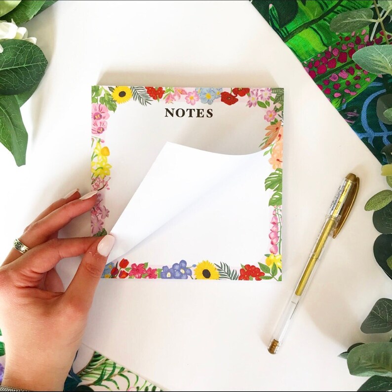 May include: A notepad with a floral border and the word "NOTES" printed in black. The notepad is white with a floral border in shades of pink, red, yellow, blue, and green. The notepad is partially open, revealing a blank white page.