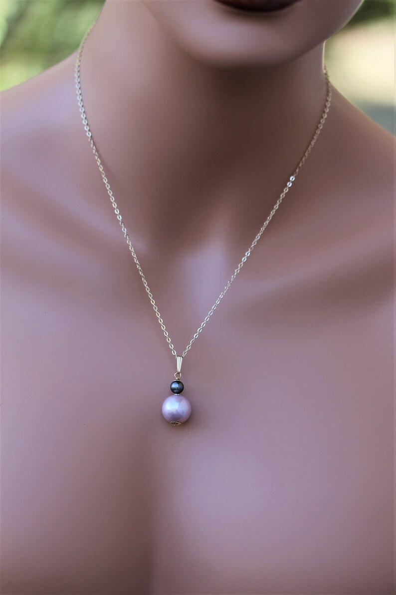 May include: A delicate gold chain necklace with a pendant featuring a single large, light pink pearl and a smaller, dark grey pearl.
