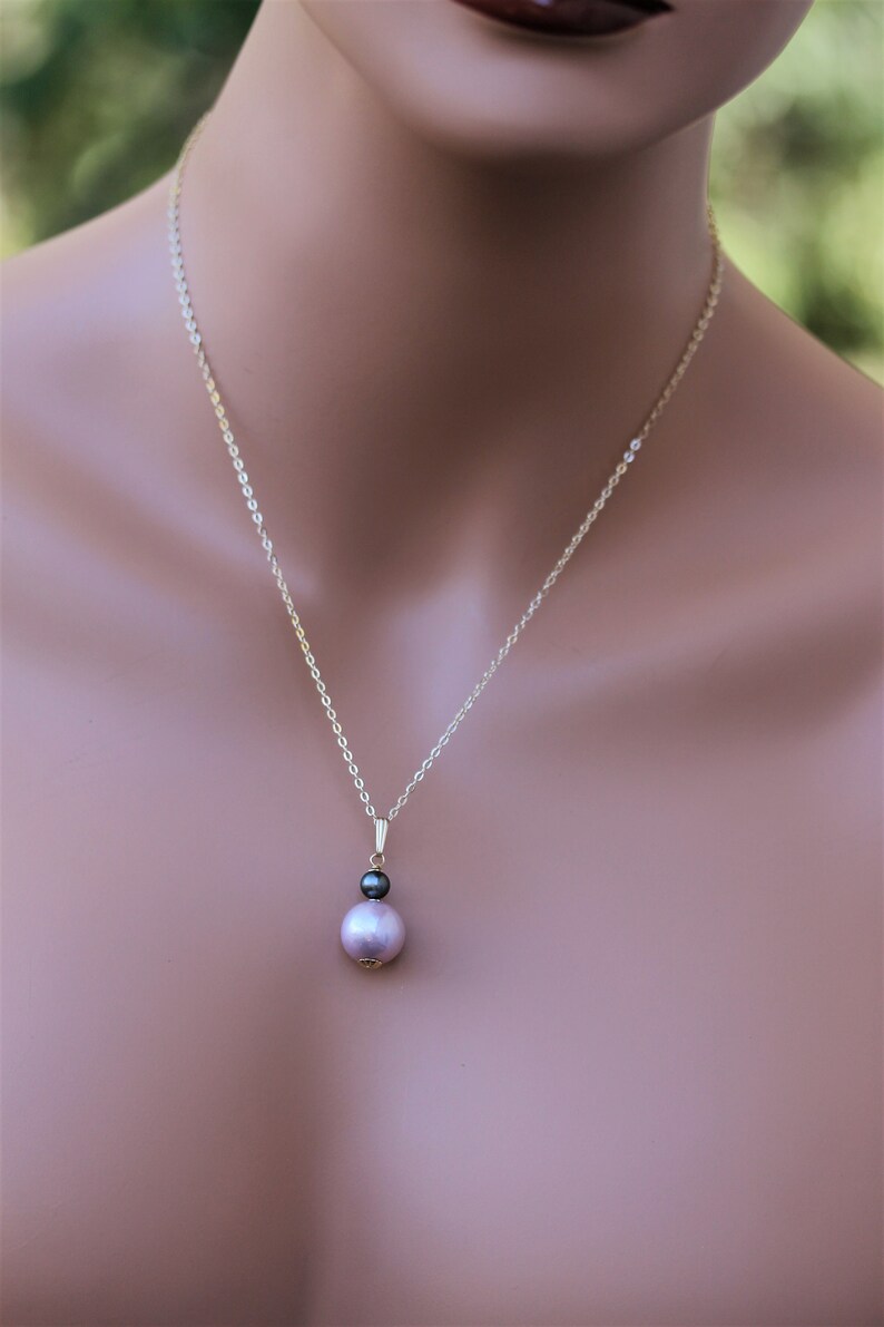 May include: A delicate gold chain necklace with a pendant featuring a large, light purple pearl and a smaller, black pearl.