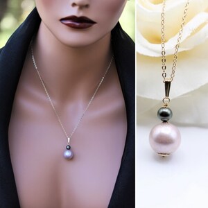 May include: A delicate gold chain necklace with a pendant featuring a large, light pink pearl and a smaller, dark gray pearl.