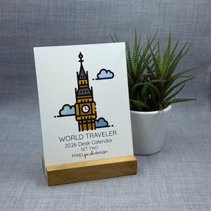 Pode incluir: Calendário de mesa "World Traveler 2026" com uma ilustração de Big Ben e nuvens azuis, em um suporte de madeira. À direita do calendário há uma pequena planta em vaso.
