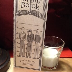 May include: A card with the text "In my Book" and a drawing of three people standing in a lineup. The text below the lineup says "You're the one who dunnit!"