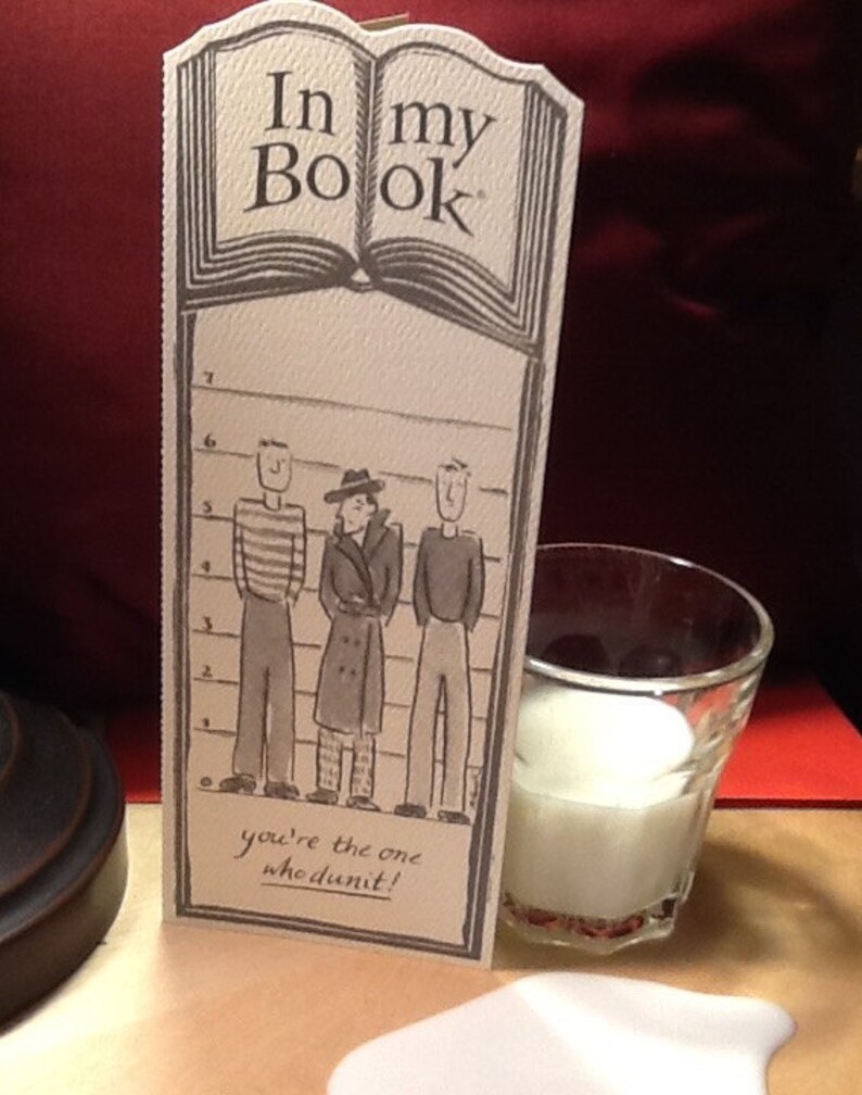 May include: A card with the text "In my Book" and a drawing of three people standing in a lineup. The text below the lineup says "You're the one who dunnit!"