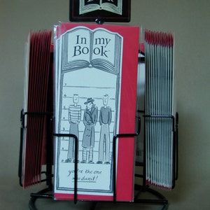 May include: A black metal display stand with red and white bookmarks. The bookmarks feature a cartoon illustration of three men in a lineup and the text "In my Book" and "you're the one whodunit!"