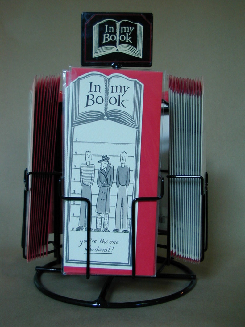 May include: A black metal display stand with red and white bookmarks. The bookmarks feature a cartoon illustration of three men in a lineup and the text "In my Book" and "you're the one whodunit!"