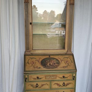 May include: An antique secretary desk with a glass-front cabinet on top. The desk is painted in a light green and gold color scheme with floral and scrollwork designs. The cabinet has a single shelf and is topped with a decorative finial.