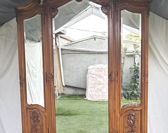 Puede incluir: Un gran armario de madera ornamentado con tres puertas con espejo. El armario es de color marrón cálido y presenta intrincadas tallas y detalles decorativos. El espejo central está enmarcado por un borde de madera detallado, y los espejos laterales son más pequeños. El armario tiene una parte superior curvada y se apoya en cuatro patas.