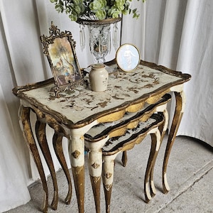 May include: A set of three antique nesting tables with ornate, curved legs. The tables have a cream-colored surface with gold accents and a decorative border. A small framed picture, a vase with greenery, and a small jar are on the top table.