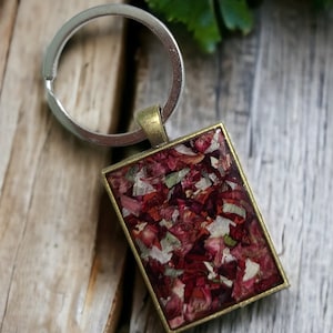 May include: A silver key chain with a square-shaped charm. The charm is filled with red, pink, and white flower petals. The charm is attached to a silver key ring.