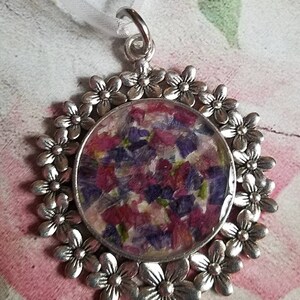 May include: A silver pendant with a round, clear resin charm filled with pressed purple and pink flowers. The pendant is surrounded by a circle of silver flower charms.