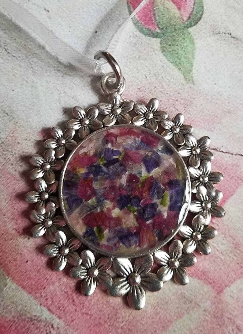May include: A silver pendant with a round, clear resin charm filled with pressed purple and pink flowers. The pendant is surrounded by a circle of silver flower charms.