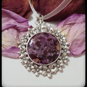 May include: A silver pendant necklace with a round, clear resin charm filled with dried purple flower petals. The charm is surrounded by a decorative silver frame with a floral design.