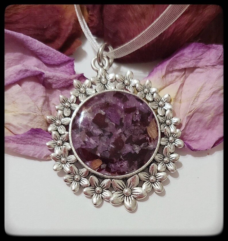 May include: A silver pendant necklace with a round, clear resin charm filled with dried purple flower petals. The charm is surrounded by a decorative silver frame with a floral design.