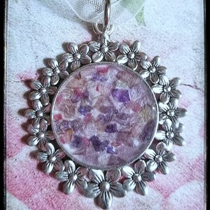 May include: A silver pendant with a round, clear resin charm filled with pressed flowers in shades of purple, pink, and white. The pendant is surrounded by a decorative border of silver flowers.