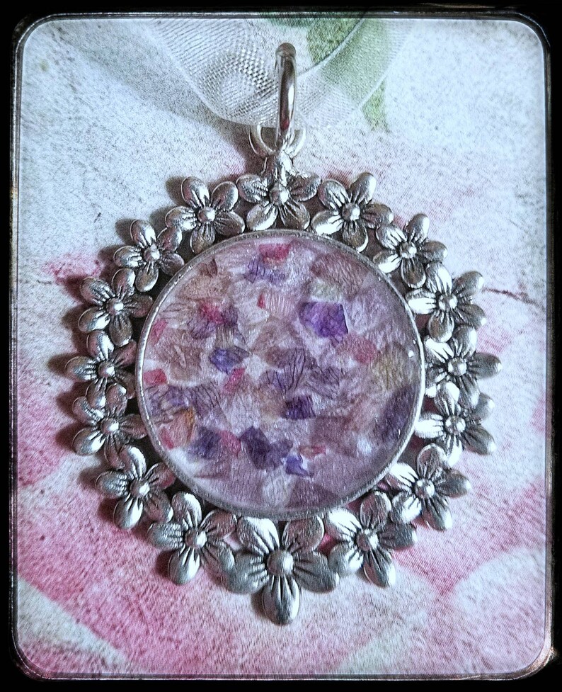 May include: A silver pendant with a round, clear resin charm filled with pressed flowers in shades of purple, pink, and white. The pendant is surrounded by a decorative border of silver flowers.