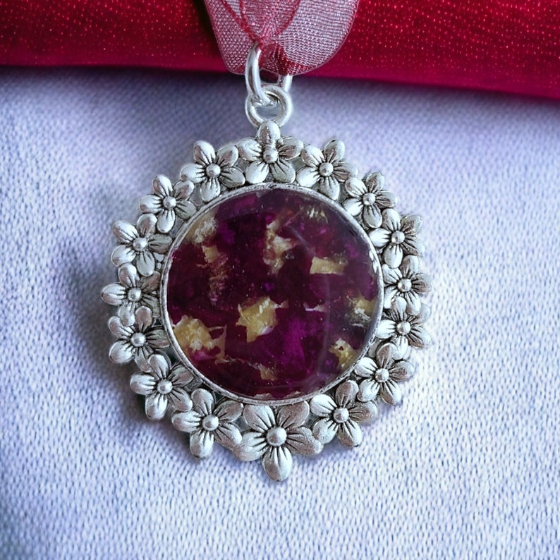 May include: A silver pendant with a circular design featuring a floral border and a center filled with pressed purple and gold flowers.