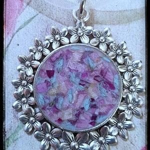 May include: A silver pendant with a round, clear resin charm filled with pink and blue flower petals. The pendant is surrounded by a circle of silver flowers.