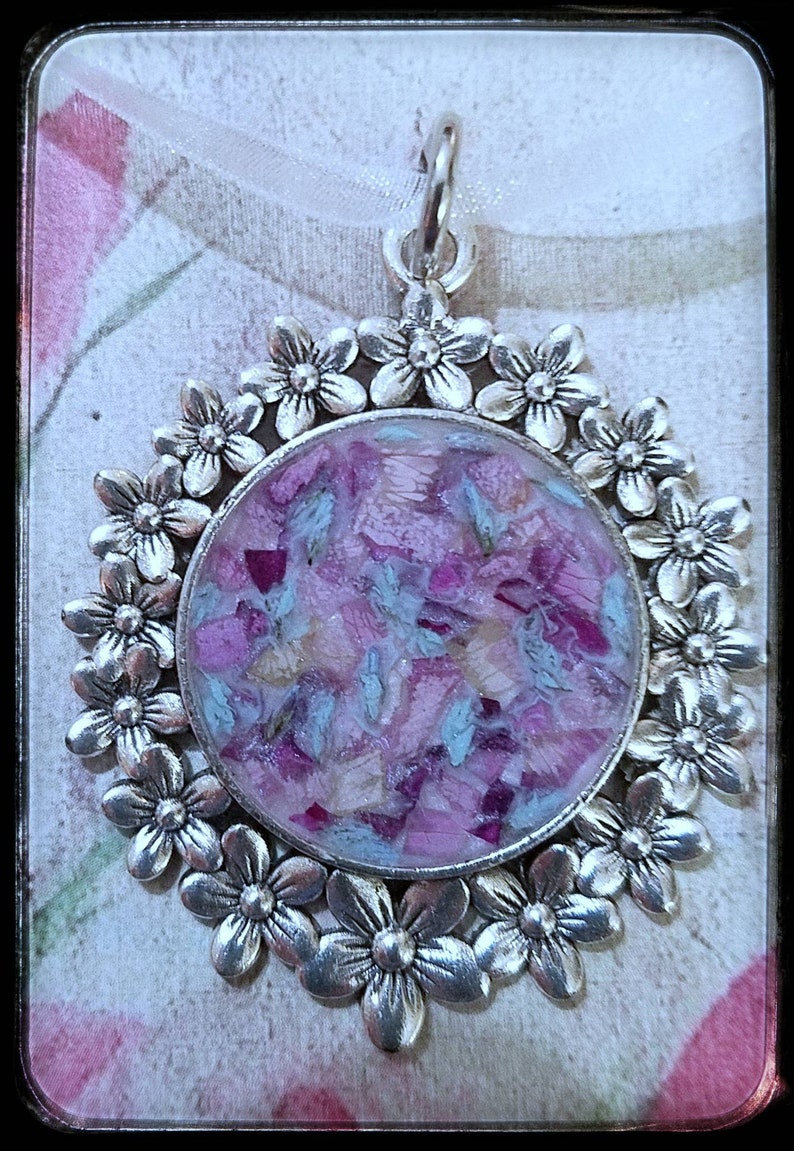 May include: A silver pendant with a round, clear resin charm filled with pink and blue flower petals. The pendant is surrounded by a circle of silver flowers.