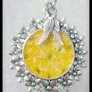 May include: A silver pendant with a round, yellow resin charm containing pressed flowers. The charm is surrounded by a silver frame with a floral design. Two silver angel wings are attached to the top of the charm.