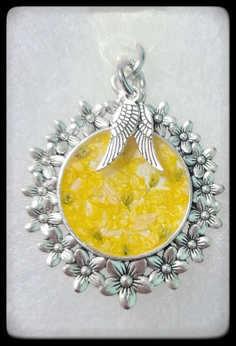May include: A silver pendant with a round, yellow resin charm containing pressed flowers. The charm is surrounded by a silver frame with a floral design. Two silver angel wings are attached to the top of the charm.