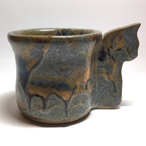 May include: A handmade ceramic mug with a unique, asymmetrical handle. The mug has a mottled glaze in shades of blue, brown, and gold.