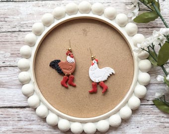 May include: A pair of dangle earrings featuring a brown and white rooster and a white hen with red boots. The earrings are displayed on a white circular background.