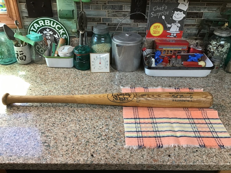 Pu&ograve; includere: Una mazza da baseball in legno Louisville Slugger con il testo "BAT 1984 CHAMPION Hall of Fame" sul fusto. La mazza &egrave; appoggiata su un panno a quadri rosa e bianco.